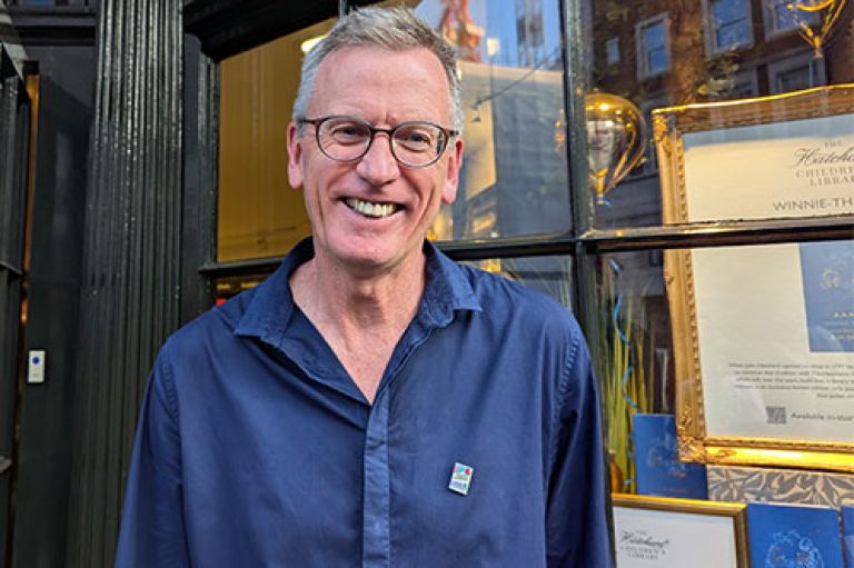 Richard Jerram wears a dark blue shirt and smiles to camera. On his shirt is a tiny pin of the cover of his book Makoto Murders. Behind him we can see the window of Hatchards in Piccadilly