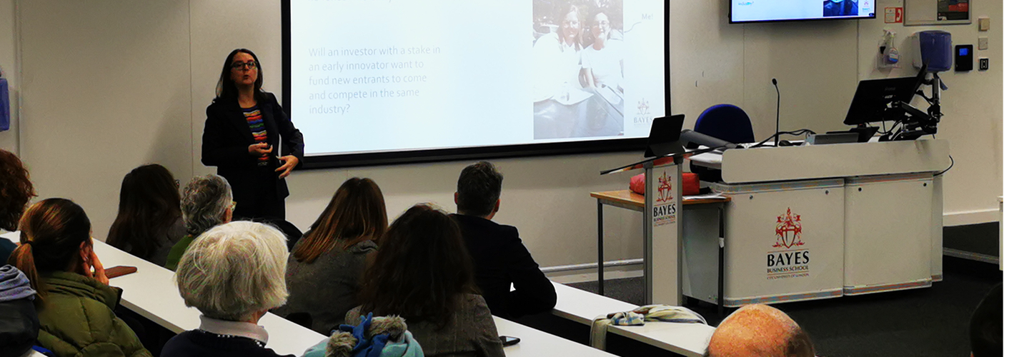 Giacinta talks while standing in front of a screen with a graph on and the image also captures the back of the heads of several audience members