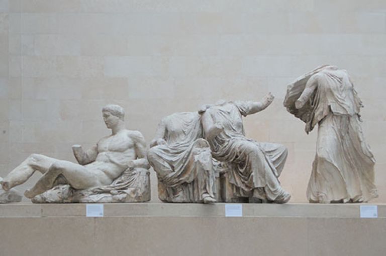 An image of a part of the Elgin Marbles. A statue of a man lays next to two seated sculpted people in flowing robes (with no heads), next to another person standing with robes flowing in air. These statues are against a pale background in the British Museum