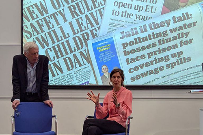 An image of Esme Wren, Editor of Channel 4 News, sitting on a chair and speaking to students. Beside her, Prof Barney Jones stands and leans his arms against a chair. Behind them shown on a screen are various newspaper with the headlines of the day – which are around fines for water bosses covering up sewage spills, and the Online Safety Act protecting children.