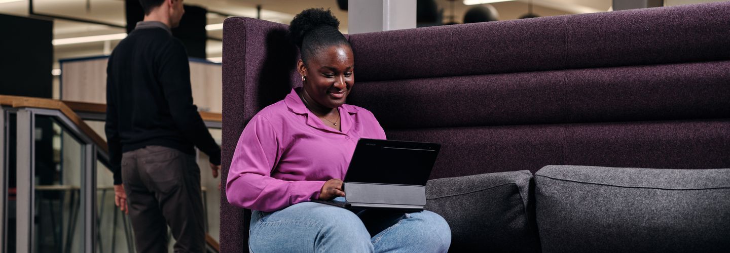A young woman sits on a purple cushioned sofa, smiling at her laptop.