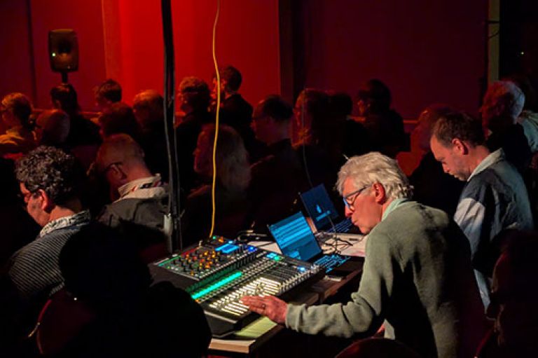 An image of Prof Simon Emmerson leaned over a mixing deck, with his hand on one of the channel faders. The light is pooled on him. To his right sits Dr Erik Dr Erik Nyström. There is a connected laptop between them and a large amp above them just out of shot. Around them, the room is dark with a faint red light and rows of people are sitting facing away from the camera (towards the stage) and listening.