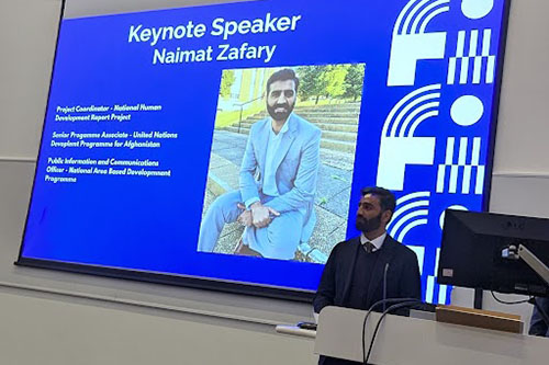 A man presents before a large digital screen displaying a slide introducing him as keynote speaker Naimat Zafahry.