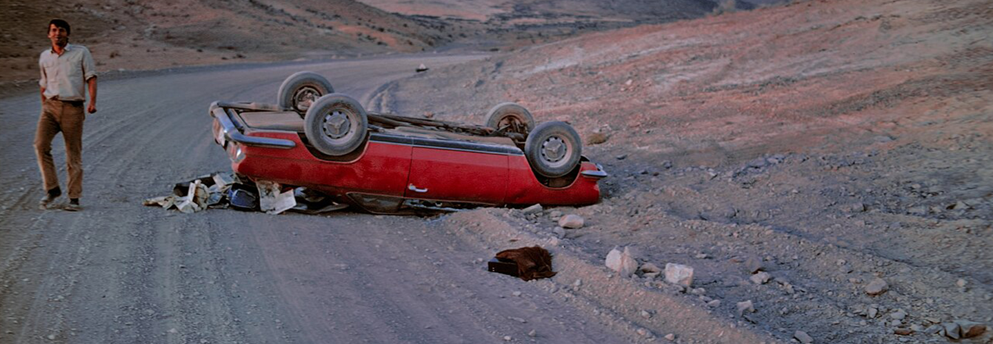 A man on a deserted road looks out at the camera standing near his overturned red car