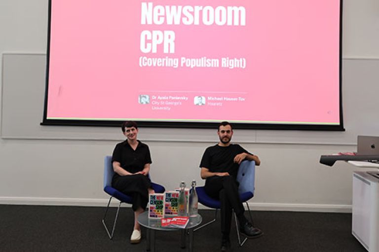 Dr Ayala Panievsky and Michael Hauser-Tov sit on blue chairs and smile to camera. In front of them is a small round coffee tables with Dr Panievsky's book 'The New Censorship.' Behind them is a big digital screen with a red background that reads 'Newsroom CPR: Covering Populism Right.'