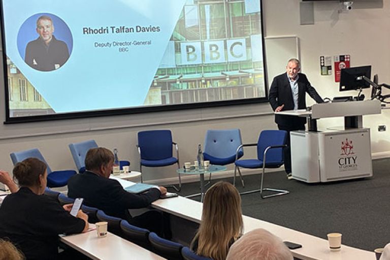 Rhodri Talfan Davies stands in front of a lectern which has the City St George's logo on it and addresses an audience in a lecture room in a horse shoe shape. In the audience can be seen Sir John Whittingdale