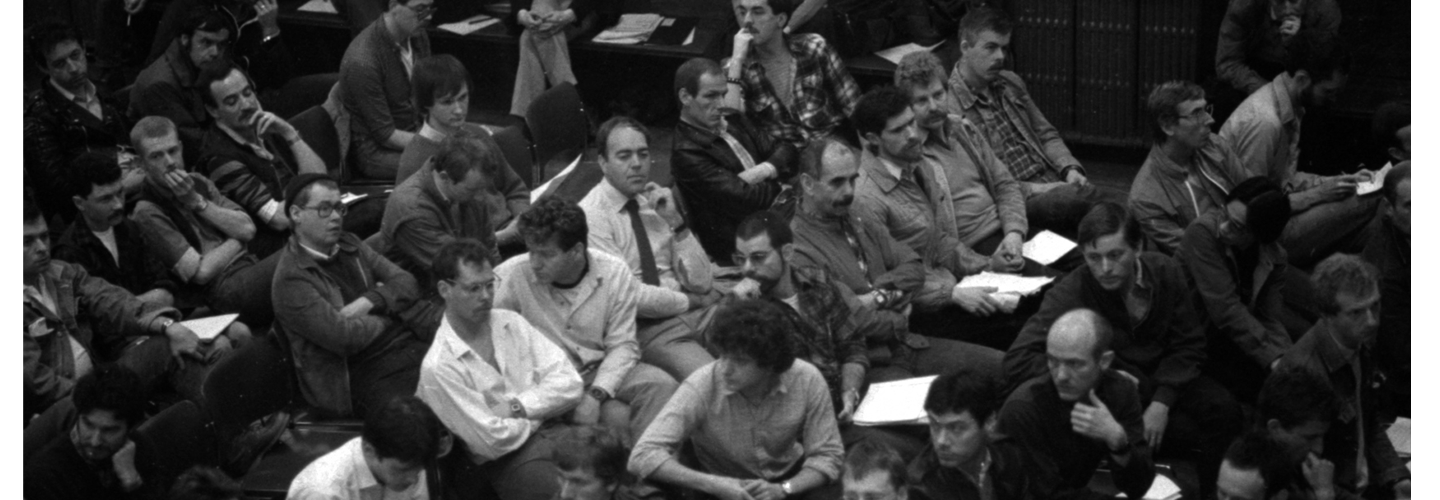 A black and white picture of several dozen people, mostly men, sitting in a public meeting hall