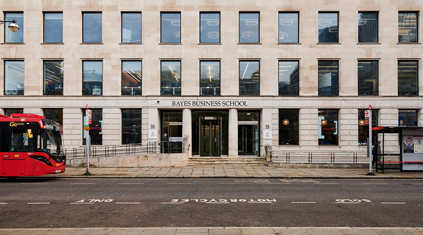 Exterior of Bayes Business School building with large glass windows and a stone facade, with a red bus and bus stop in front.