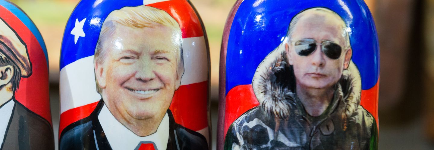 An image of Russian dolls, one depicting a photo of Trump and the other of Putin, with their respective countries' flags as a background.