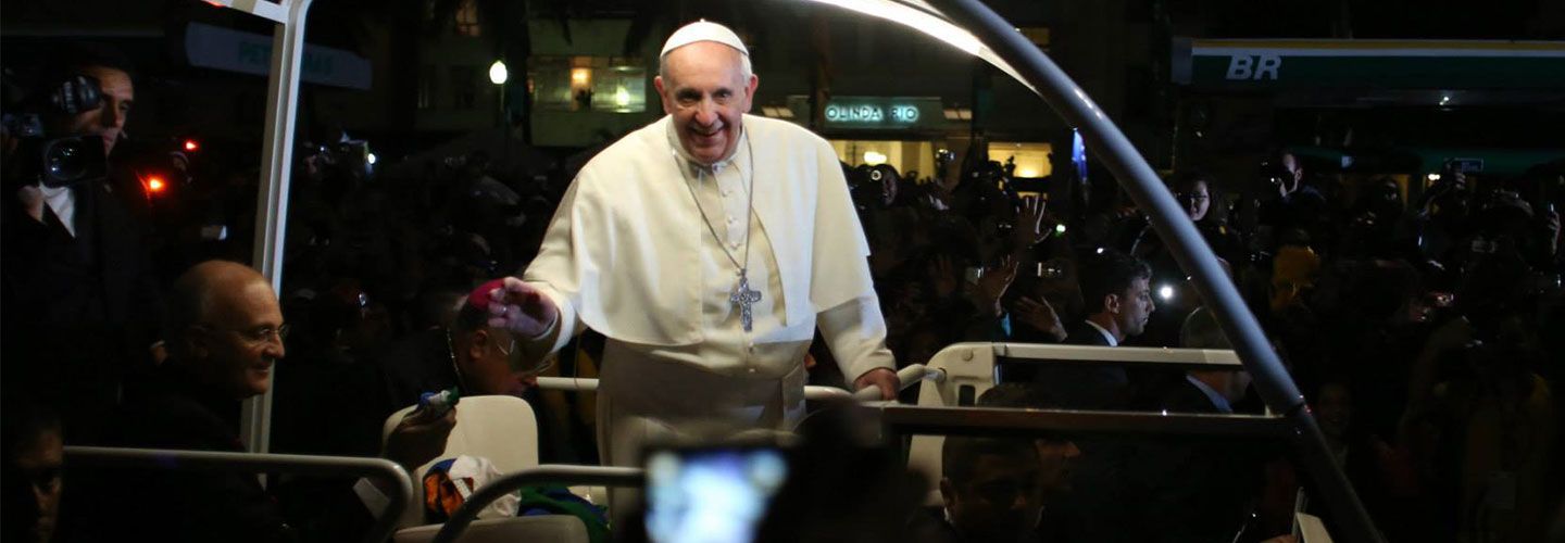 An image of Pope Francis wearing all white and a long silver cross necklace, smiling to camera. He is in an open vehicle with lights above his head. The background around him is dark but you can perceive crowds of people and photographers