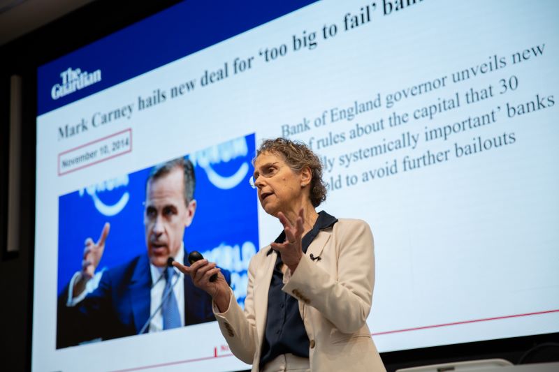 A woman makes a point on the stage with a picture of Mark Carney behind her