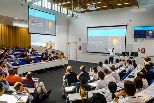 A man in front of an illuminated screen is talking to a conference room where about 30 people are visible