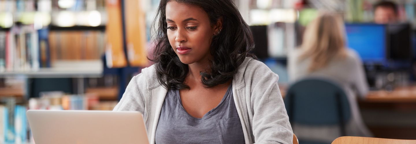 A young woman sits in a library, focused on her laptop.
