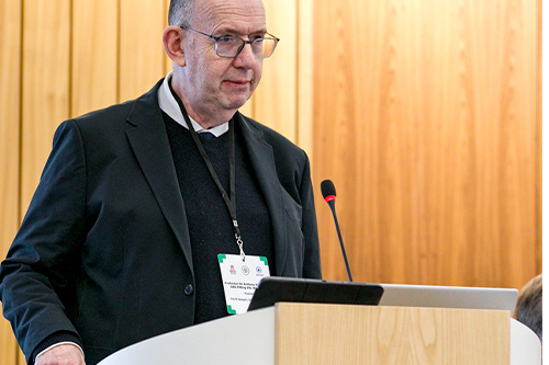 A man stands at a lectern with the Bayes logo with a picture of men in 1970s fashion in the background