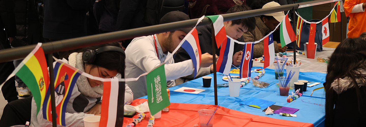 A group of students sit at a table painting flags, flags from the world hang above them