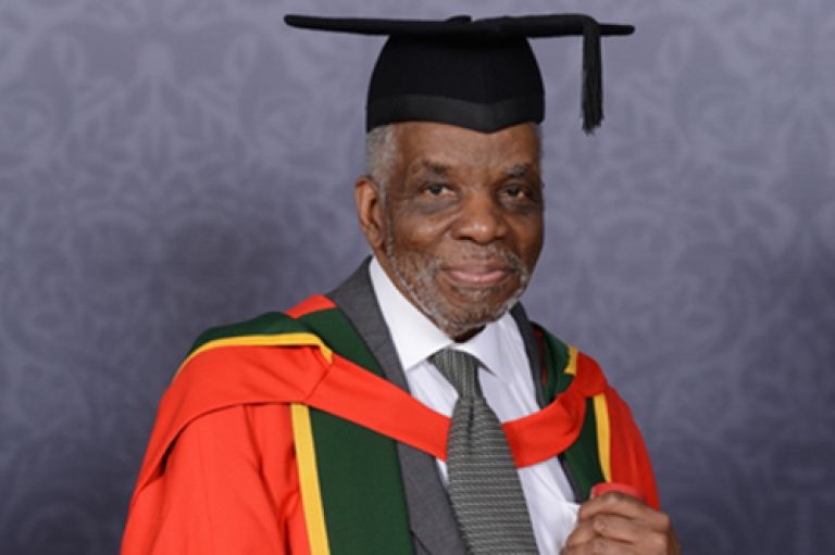 Aggrey Burke, wearing ceremonial honorary graduation robes and cap, holding scroll whilst smiling into camera.