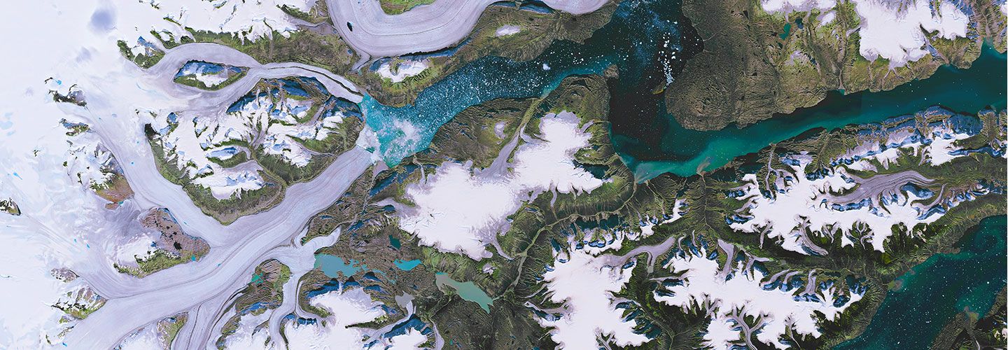 An aerial shot of Scoresby Sund, which is the longest fjord in the world, stretching for 350 km (217 miles), and also one of the deepest (over 1500 m, 4,900 ft deep). Waters in the fjords show spots of bright turquoise color from sediments deposited there by runoff. Little spots of white in the water seem to be ice originating from the deeper fjords that reach all the way to the icecap covering most of the island.