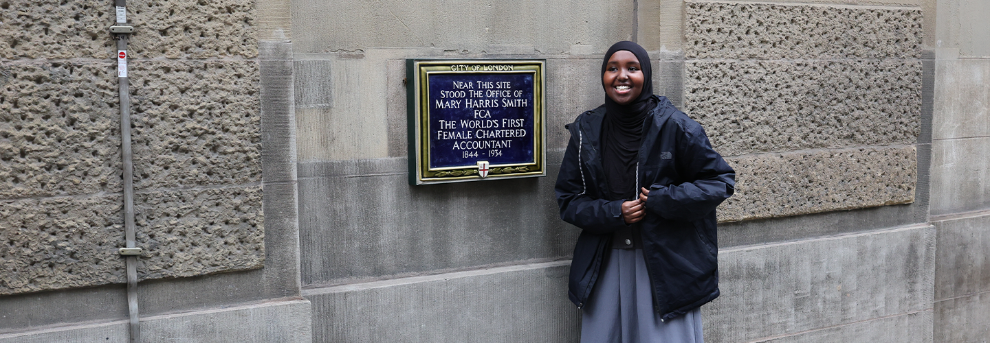 A young woman stands next to a blue plaque marking the place where Mary Harris Smith, the world's first female chartered accountant, had her office.