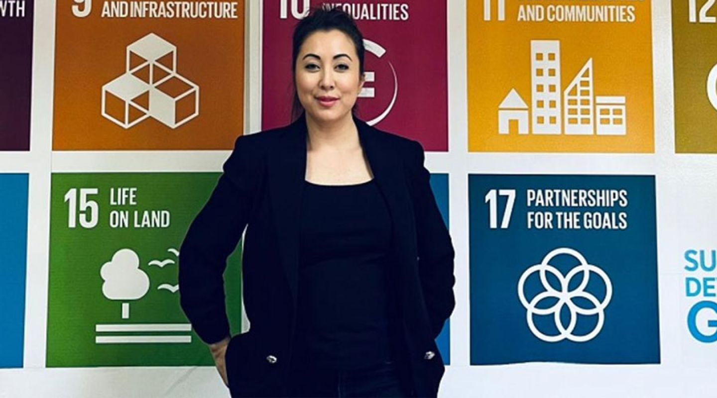 Dr Sophie Knowles smiles to camera, standing in front of a banner of the UN global goals. They are brightly coloured tiles and with little icons and words and numbers, such as 8 Decent Working and Economic Growth, and 14 Life Below water.
