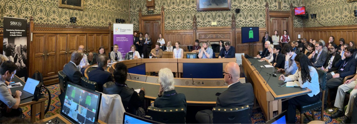An image of speakers at the event in a room in the House of Commons. We can see the round wooden table with speakers along it with small microphones in front of them. Behind them , above wooden panelling, are very intricate wallpaper designs and an old portrait oil painting. On either side of the horseshoe desk are banners, one for SOUK and one for the Foreign Policy Centre