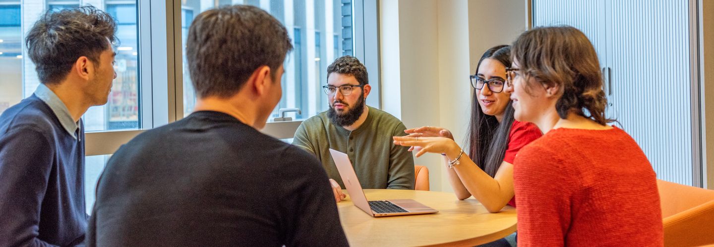 Mixed group of students sat around a table with laptop, having a discussion.