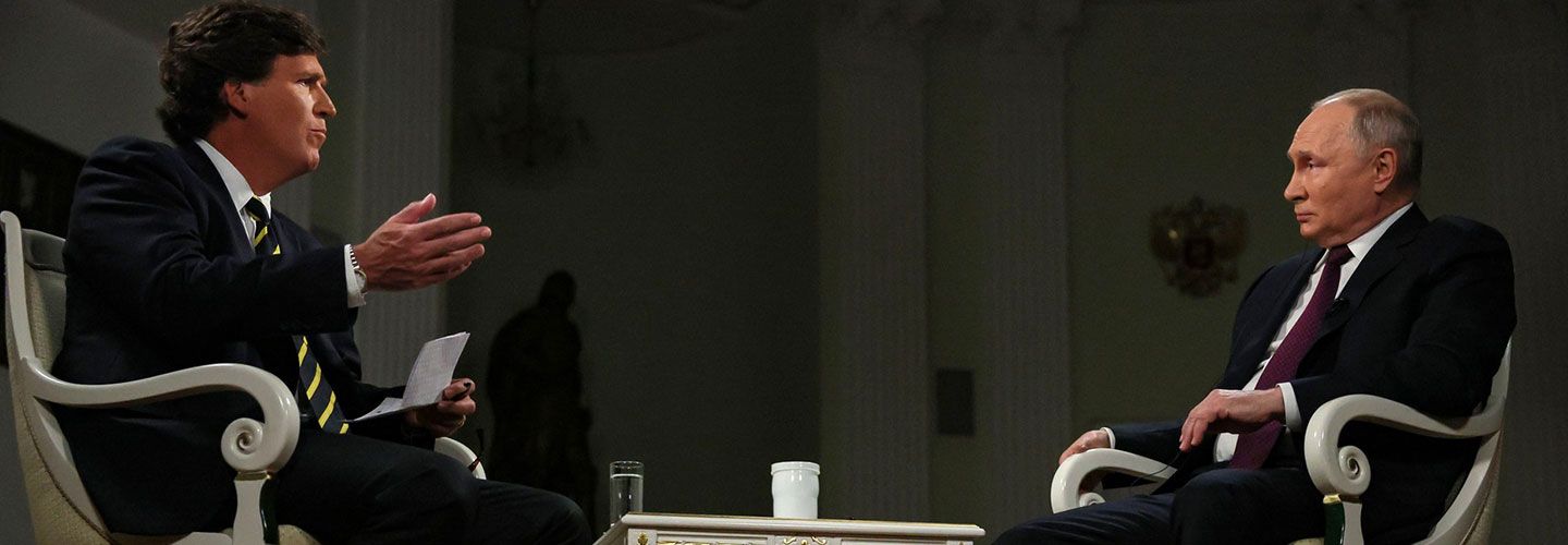 Image of Tucker Carlson interviewing Vladimir Putin. Both sit on wooden side chairs with a smalll white wooden table between them. Tucker holds a notepad and his hand in the air, speaking. Putin is leaning to one side and listening.