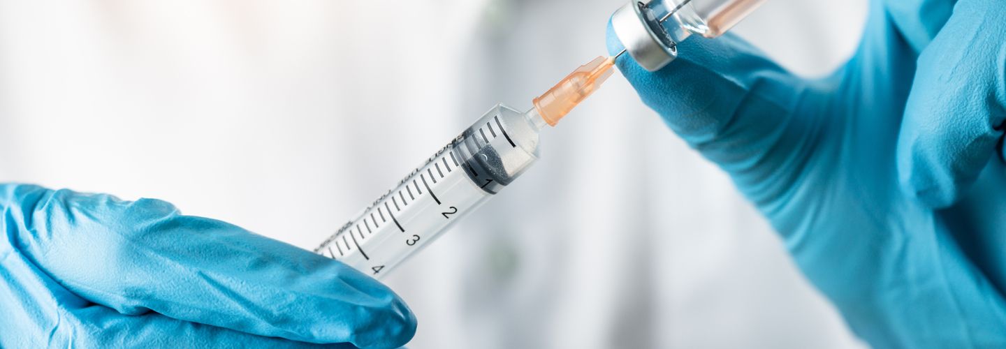 An image of two hands in blue gloves holding a vial of liquid (a vaccine) in one hand and and needle in the other 