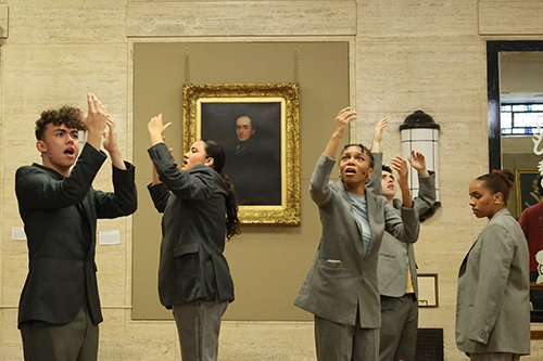 Performers wearing suits stare intently at hands as they hold them in the air