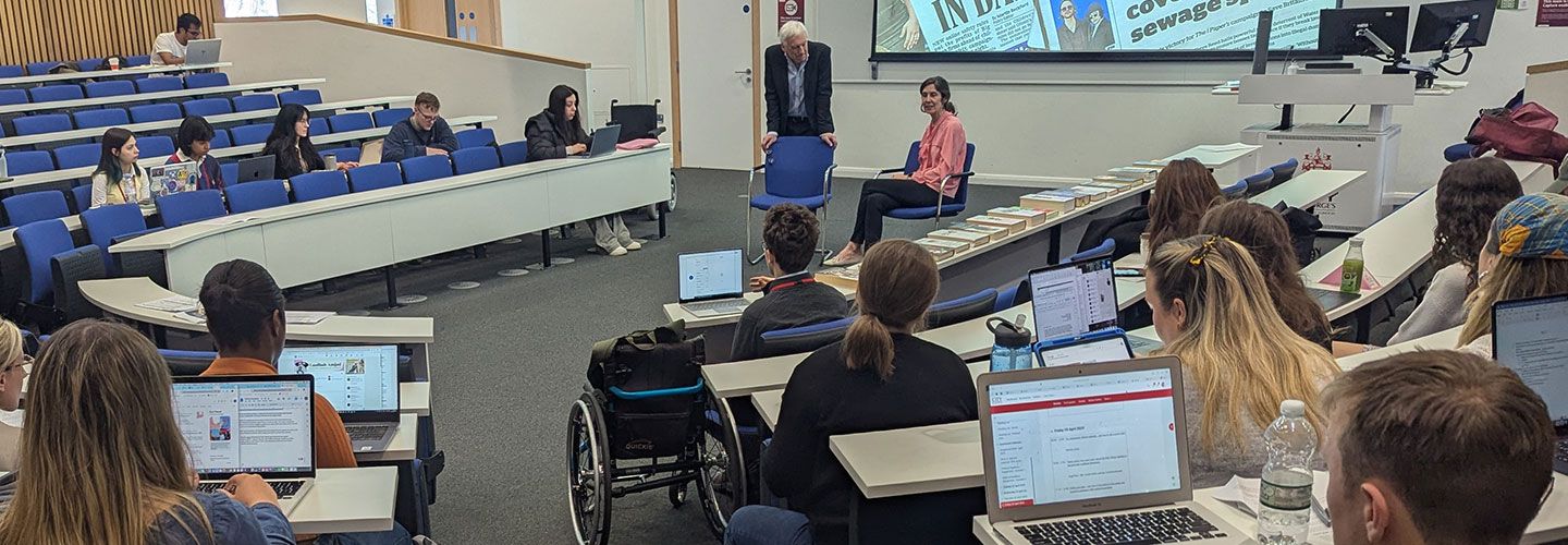 An image of Esme Wren, Editor of Channel 4 News, sitting on a chair and speaking to students. Beside her, Prof Barney Jones stands and leans his arms against a chair. Behind them shown on a screen are various newspaper with the headlines of the day – which are around fines for water bosses covering up sewage spills, and the Online Safety Act protecting children.