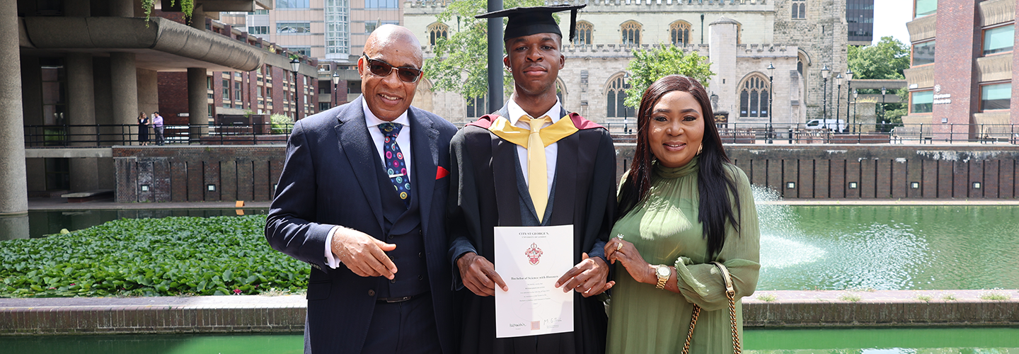 A young man in a cap and gown holds his certificate with his father (wearing a suit) to his left and his mother (wearing a green dress) to his right