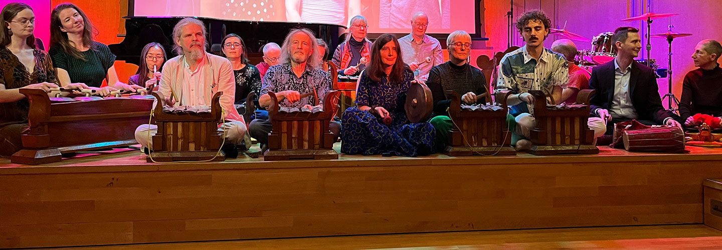 A row of people sit on stage with gamelans in front of them. They are about to perform