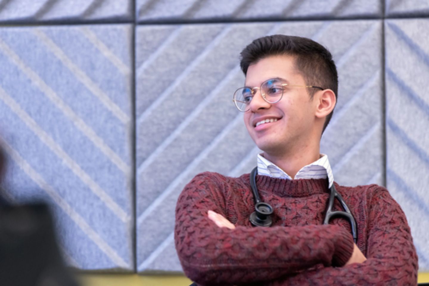 Smiling person with a stethoscope around their neck, wearing glasses and a red sweater, sits against a patterned gray wall, conveying a relaxed mood.