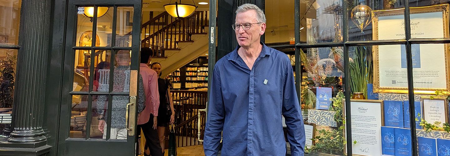 Richard Jerram wears a dark blue shirt and looks to the left. On his shirt is a tiny pin of the cover of his book Makoto Murders. Behind him we can see the window of Hatchards in Piccadilly and its door, with people mingling inside buying copies of his book