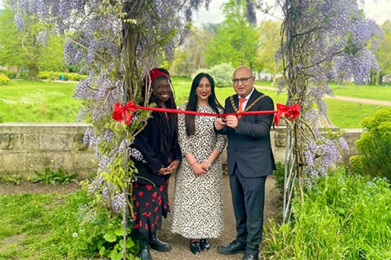 Three people stand under a wooden arch with wisteria growing on it. At their shoulders is a red sash. The Mayor of Ealing holds scissors to cut this. They stand in a green park.