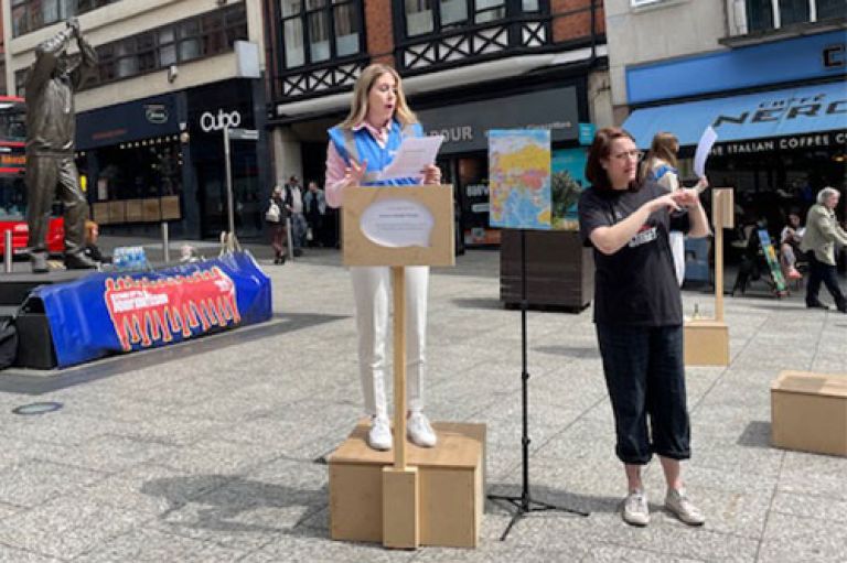A student journalist stands on a small wooden crate which acts as a podium to deliver news. She stands in a main square in Nottingham city centre. Next to her stands a sign language interpreter