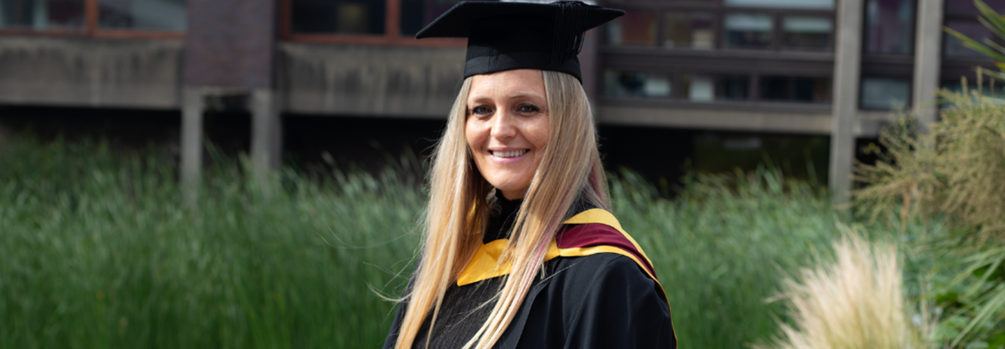 A woman in a cap and gown is outside with trees behind