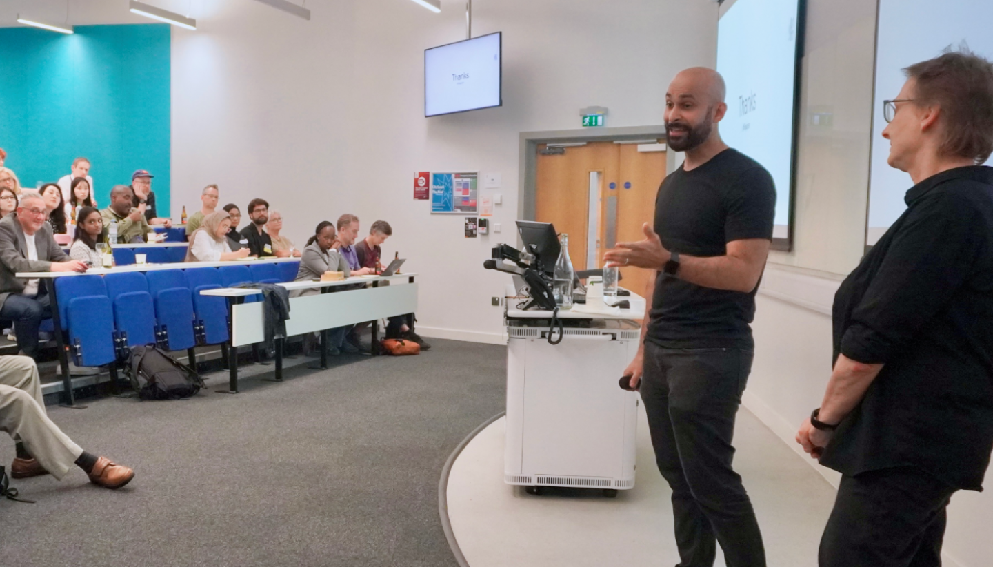 Raj Arjan, Project Manager at Google, is seen presenting, gesturing at the audience. Standing next to him is Professor Stephanie Wilson