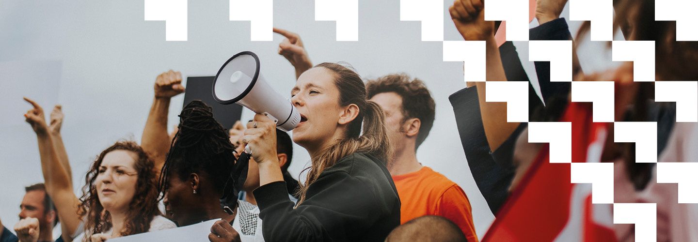 Crowd of people with arms raised in a rally. A woman in the foreground shouts through a loudspeaker.