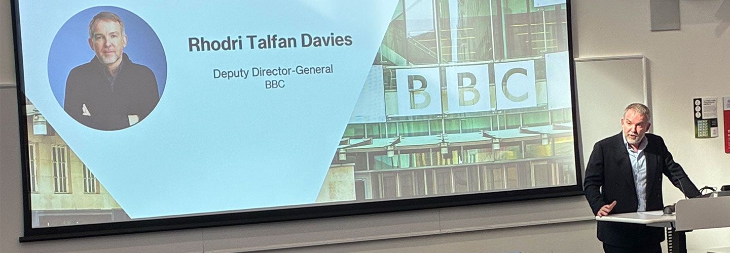 Rhodri Talfan Davies stands in front of a lectern which has the City St George's logo on it and addresses an audience in a lecture room in a horse shoe shape. In the audience can be seen Sir John Whittingdale