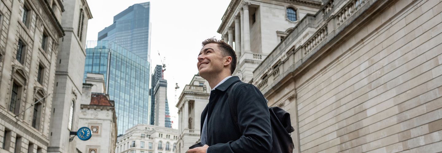 A City St George's student dressed smartly in a long winter jacket and a white shirt smiles and looks upwards as he crosses Fenchurch Street. To his right are the walls that lead to the Bank of England, and skyscrapers can be send in the distance behind him