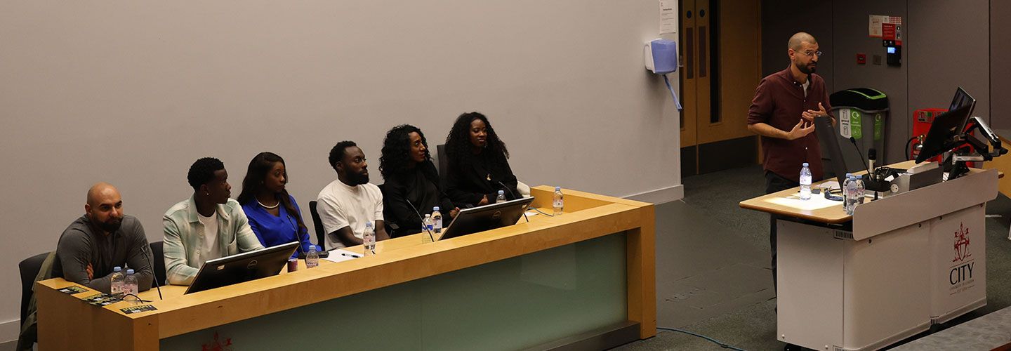 The speakers sit in a row at a desk with small microphones in front of them. To the right, Lambros gestures and speaks to an audience
