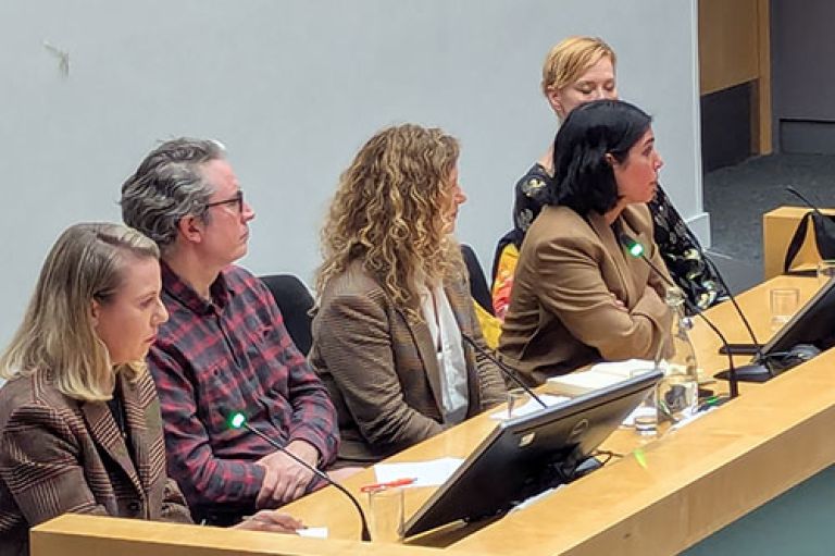 An image of the speakers sitting at a desk in a row. The desk is a light beige colour and the image is cropped closely to them. Bent slightly forward and addressing the audience is Mirren Gidda, barrister and former journalist