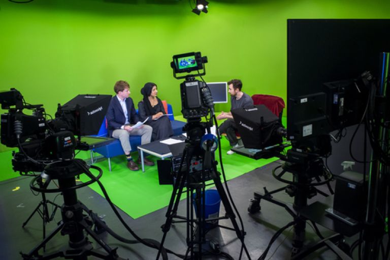 Three people sit on chairs against a green screen, with notes on their laps, appearing to film a broadcast news segment. To the foreground of the image is professional news camera equipment recording